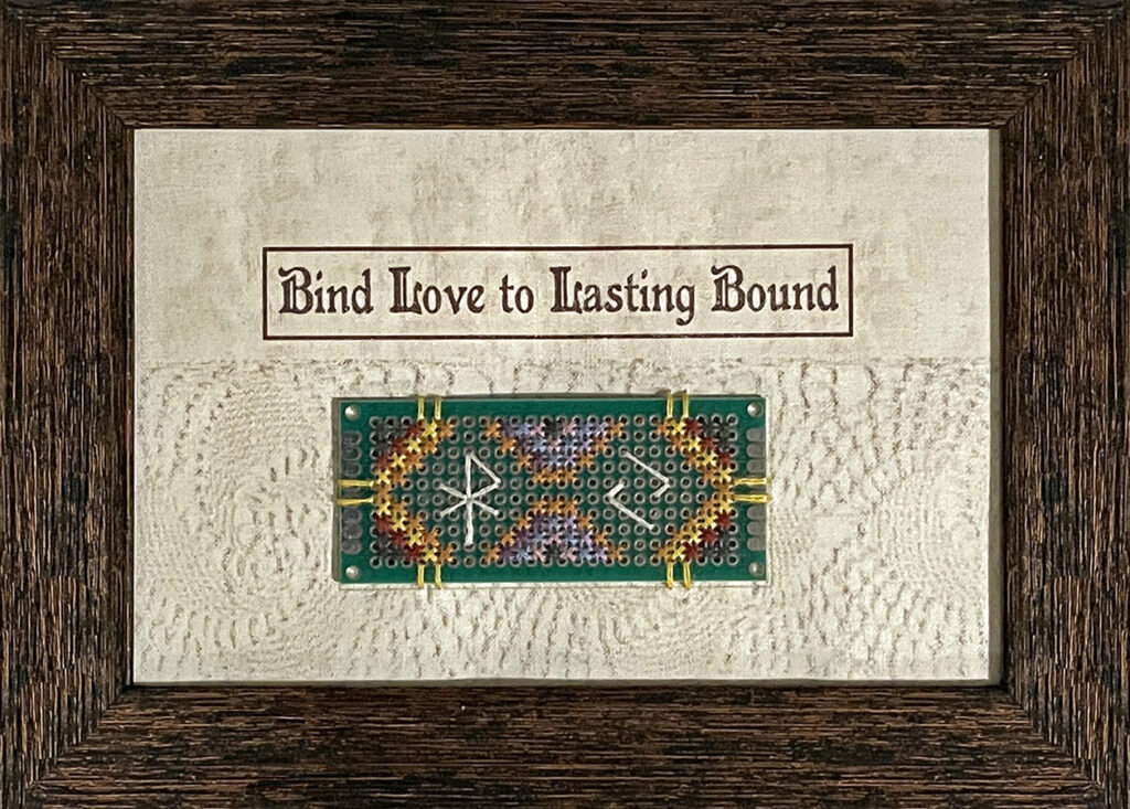 A framed piece of cream-colored fabric printed with the words "Bind Love to Lasting Bound". Below the words is an embroidered design on a small rectangular circuit board, sewn on a strip of lace.